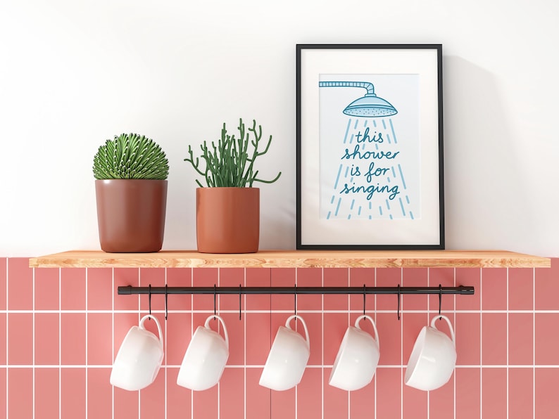 May include: A black-framed print with the text "this shower is for singing" is displayed above a shelf with two potted plants and five white mugs. The print features a blue showerhead illustration. The shelf is above a pink tiled wall.