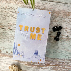 Könnte beinhalten: Ein Buch mit dem Titel "Trust Me" von Jennifer Jancke, mit goldfarbenen Buchstaben auf einem Cover im Aquarellstil. Das Cover zeigt eine Stadtlandschaft und goldfarbene Akzente. Zwei schwarze Schachfiguren und eine Muschel sind ebenfalls vorhanden.