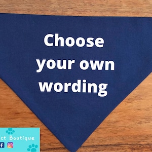 以下が含まれることがあります： 中央に白い文字で「Choose your own wording」とプリントされた、三角形のネイビーブルーのペット用バンダナ。このアクセサリーは端が丁寧に仕上げられており、平らに置かれた状態で、無地の濃い青色と太い文字が際立っています。
