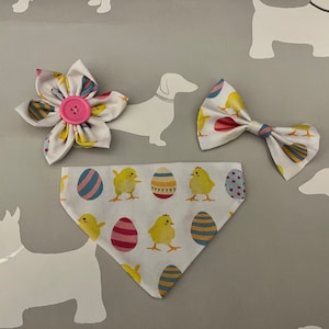 May include: A collection of Easter-themed pet accessories on a grey backdrop. The set includes a flower with a pink button, a bow tie, and a bandana, all patterned with Easter eggs and yellow chicks. White dog silhouettes are scattered around.