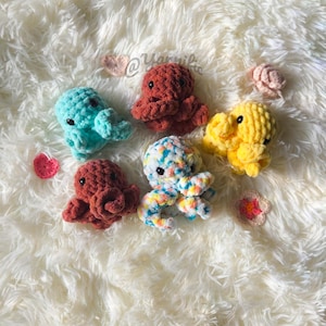 May include: A collection of six crocheted octopus toys in various colors, including teal, rust, yellow, and a multicolored option. The toys are arranged on a fluffy white surface, with small crocheted flowers scattered around.