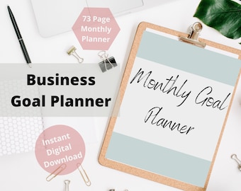 May include: A white clipboard with a light blue background and the text "Monthly Goal Planner" written in black cursive. The clipboard is on a white surface with a laptop, a pen, and paper clips.