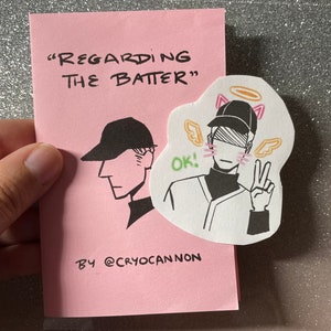 Puede incluir: Una tarjeta rosa con el texto "Regarding the Batter" y una ilustración en blanco y negro de una persona con una gorra de béisbol y una camiseta. La persona está levantando dos dedos y tiene un halo y orejas de gato. La tarjeta también tiene el texto "By @cryocannon" en la parte inferior.