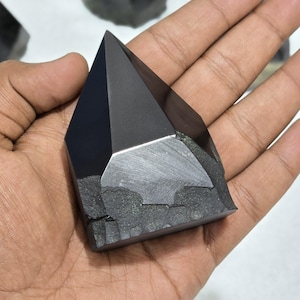 May include: A black obsidian crystal point with a polished, faceted top and a rough base. The crystal is held in a hand, highlighting its dark, reflective surface. Other similar crystals are visible in the background.