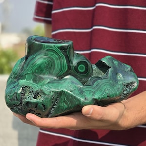 May include: A large, polished malachite stone with swirling patterns of dark and light green. The stone has a rough, natural shape with a cut section on top. The malachite is held in a person's hands.