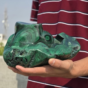 May include: A large, polished malachite stone with swirling patterns of dark and light green. The stone has a unique, organic shape with a carved-out section and a smooth, glossy surface. The malachite is held in a person's hand.