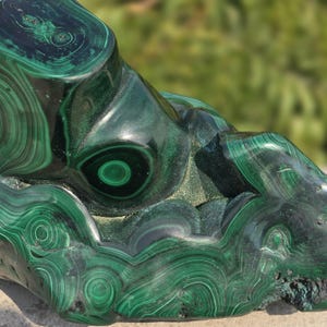 May include: A polished malachite stone with swirling patterns of dark and light green. The natural stone has a smooth, glossy surface with concentric circles and bands, showcasing its unique mineral formations. The stone's surface is partially in focus, with a blurred green background.