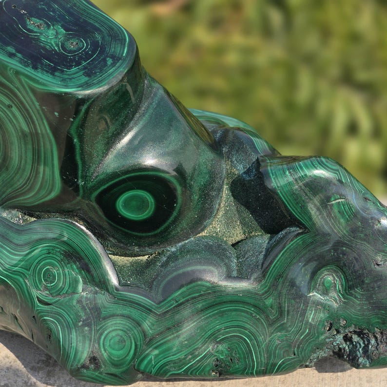 May include: A polished malachite stone with swirling patterns of dark and light green. The natural stone has a smooth, glossy surface with concentric circles and bands, showcasing its unique mineral formations. The stone's surface is partially in focus, with a blurred green background.