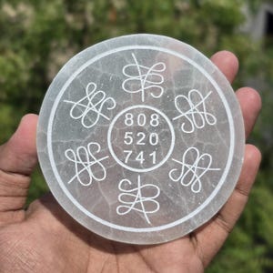 May include: A round, translucent, white stone disc with etched symbols and numbers. The center circle contains the numbers 808, 520, and 741. Six stylized symbols are arranged around the disc's edge. The disc is held in a hand.