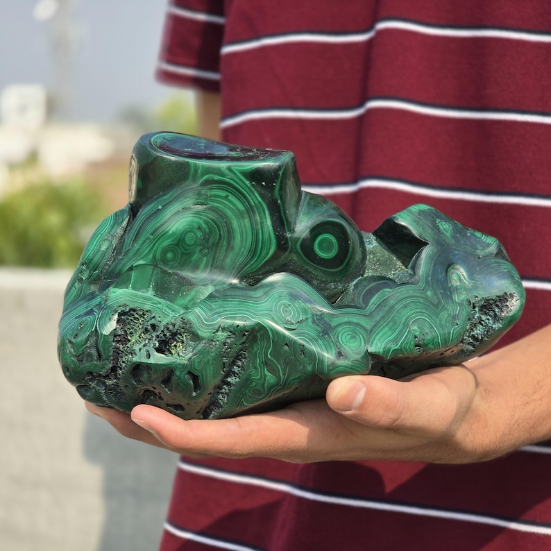 May include: A large, polished malachite stone with swirling patterns of dark and light green. The stone has a rough, natural shape with a cut section on top. The malachite is held in a person's hands.