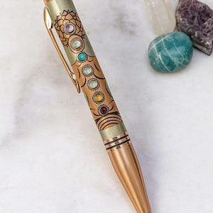 May include: A decorative pen with a copper-colored barrel and a clear top filled with glitter. The pen features a series of colorful gemstones and an ornate design along the barrel. Additional gemstones are visible in the background.