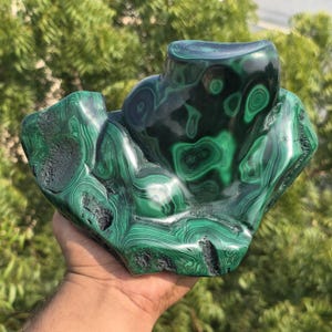 May include: A polished malachite stone sculpture with a swirling pattern of dark and light green. The stone has a unique, organic shape with a smooth, glossy surface. The sculpture is held in a hand, with a blurred green background.