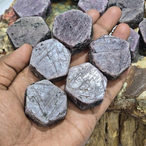 Puede incluir: Primer plano de varias piedras preciosas de rubí en bruto, de forma hexagonal. Las gemas son de un color rojo púrpura intenso, con inclusiones naturales y texturas superficiales visibles. Se sostienen en una mano.
