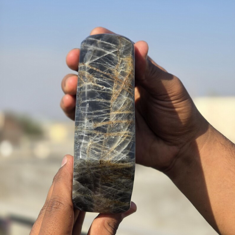 Puede incluir: Una piedra preciosa rectangular pulida con una parte superior redondeada. La piedra es principalmente de color gris oscuro con intrincadas vetas de color claro. La piedra preciosa se sostiene en las manos de una persona contra un cielo azul claro. La piedra mide unos 15 cm de largo.