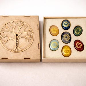 May include: A wooden box with a laser-cut tree design and a set of seven chakra stones. The oval stones feature gold chakra symbols on coloured backgrounds, including blue, green, and red. The box is open.