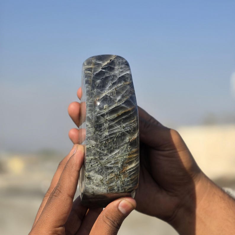 Puede incluir: Una piedra rectangular pulida con la parte superior redondeada. La piedra tiene un color base gris oscuro con intrincados patrones de vetas grises claras y marrones. La piedra se sostiene con dos manos sobre un fondo de cielo azul.