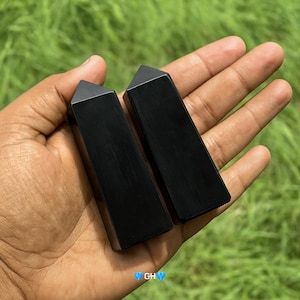 May include: Two polished black obsidian obelisks with a triangular top. The obelisks are rectangular and held in a hand against a green background. The obelisks are smooth and reflect light.