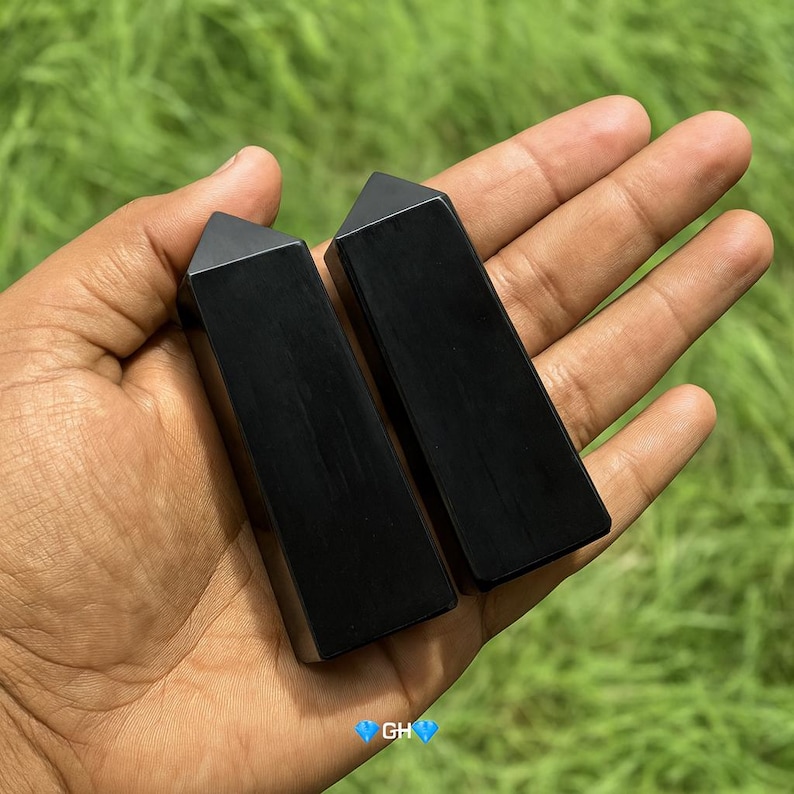 May include: Two polished black obsidian obelisks with a triangular top. The obelisks are rectangular and held in a hand against a green background. The obelisks are smooth and reflect light.
