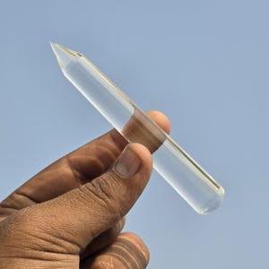 May include: A clear, transparent crystal wand with a pointed tip. The crystal is held against a blue sky background. The wand is cylindrical with a flat base and is approximately 15 cm long.