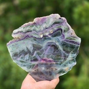 May include: A translucent, flat-cut slab of fluorite mineral featuring distinct, jagged bands of deep purple, light green, and clear white. The stone displays a natural, irregular geometric pattern with visible internal fractures and crystalline layers.