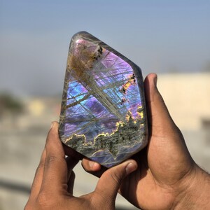 Puede incluir: Una piedra preciosa de labradorita pulida con destellos iridiscentes azules, morados y dorados. La piedra se sostiene con dos manos, mostrando su juego de colores &uacute;nico y sus patrones naturales. La piedra tiene una superficie lisa y pulida.