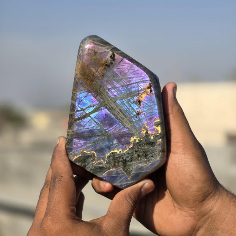 Puede incluir: Una piedra preciosa de labradorita pulida con destellos iridiscentes azules, morados y dorados. La piedra se sostiene con dos manos, mostrando su juego de colores &uacute;nico y sus patrones naturales. La piedra tiene una superficie lisa y pulida.
