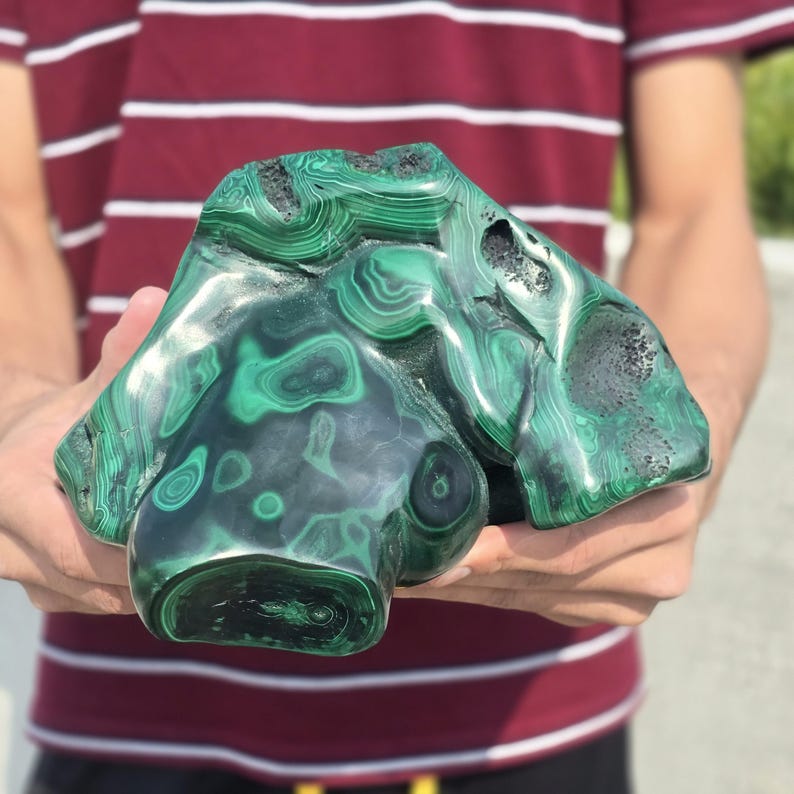 May include: A polished malachite stone with swirling patterns of dark and light green. The stone has a unique, organic shape with a smooth, glossy surface. The malachite is held in both hands.