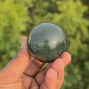Könnte beinhalten: Eine dunkelgrüne, polierte Kugel. Die glatte Oberfläche reflektiert das Licht und erzeugt Glanzlichter. Die Kugel wird in einer Hand gehalten, mit einem verschwommenen grünen Hintergrund.