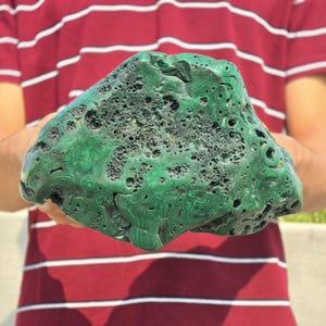 May include: A large, irregularly shaped malachite stone with a vibrant green color and intricate swirling patterns. The stone has a porous texture with numerous small holes and a rough surface. The malachite is held against a background of a red and white striped shirt.
