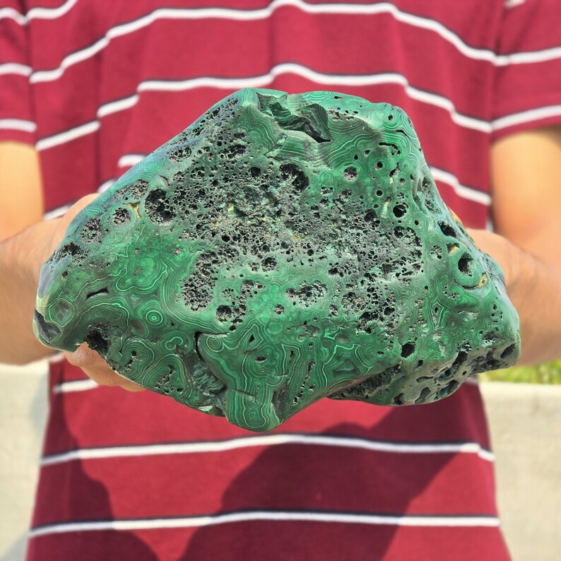 May include: A large, irregularly shaped malachite stone with a vibrant green color and intricate swirling patterns. The stone has a porous texture with numerous small holes and a rough surface. The malachite is held against a background of a red and white striped shirt.