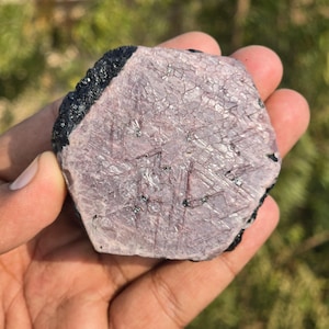 Könnte beinhalten: Ein grob geschnittener, sechseckiger Rubin-Edelstein mit einer strukturierten, hellvioletten Oberfläche und dunkel schwarzen Kanten. Der Rubin wird in einer Hand gehalten und zeigt seine natürliche, rohe Form. Der Stein hat sichtbare innere Muster.