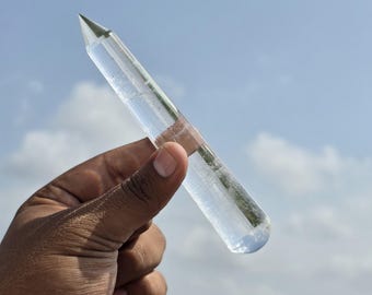 以下が含まれることがあります： 磨かれた円筒形のボディとファセット加工された尖った先端を持つ、透明なクォーツクリスタルのポイント。クリスタルは無色で、内部に自然なインクルージョンがあり、曇り空の青い背景に対して光を反射しています。