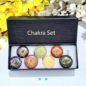 May include: A black box set labelled "Chakra Set" contains seven round stones in various colours, each with a gold chakra symbol. The stones are arranged in a row inside the box, which is lined with white fabric. The background features yellow and white flowers.