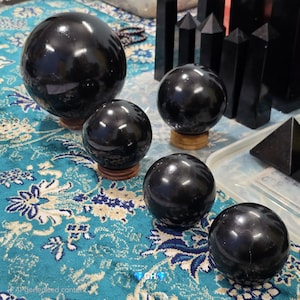 Può includere: Diverse sfere di ossidiana nera lucida di varie dimensioni sono esposte su una superficie blu e bianca a motivi. Le sfere poggiano su piccoli supporti di legno. Sono visibili anche altre forme di cristallo nero, tra cui obelischi e una piramide.