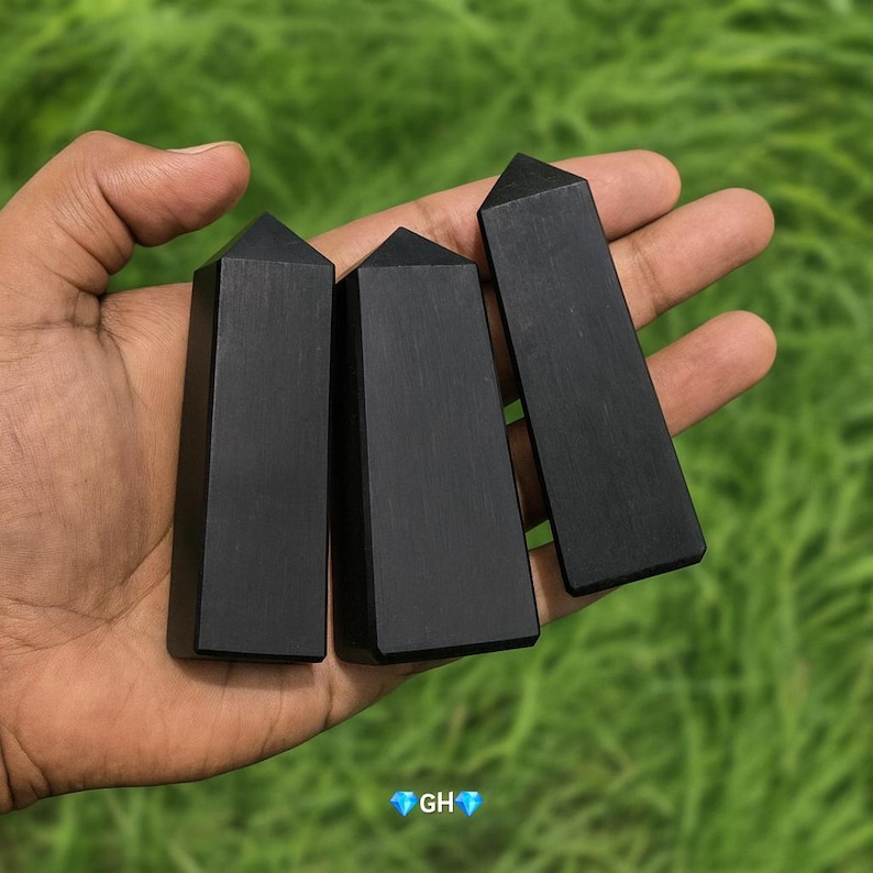 May include: Three black obsidian obelisks with a polished finish are held in a hand against a blurred green background. Each obelisk has a rectangular body and a pointed top. The obelisks are of varying lengths.