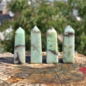Pode incluir: Quatro obeliscos de cristal verde e branco com veios castanhos são exibidos numa superfície de madeira. Os obeliscos são retangulares com pontas. O fundo está desfocado, mostrando folhagem verde.