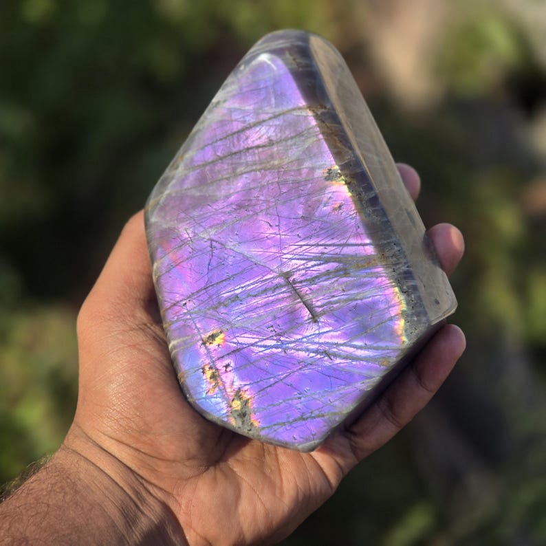 Puede incluir: Un gran cristal de labradorita pulida con una vibrante exhibici&oacute;n de colores p&uacute;rpuras e iridiscentes. La piedra se sostiene en una mano, mostrando su superficie lisa y reflectante y sus patrones naturales. La forma del cristal es aproximadamente rectangular con bordes redondeados.