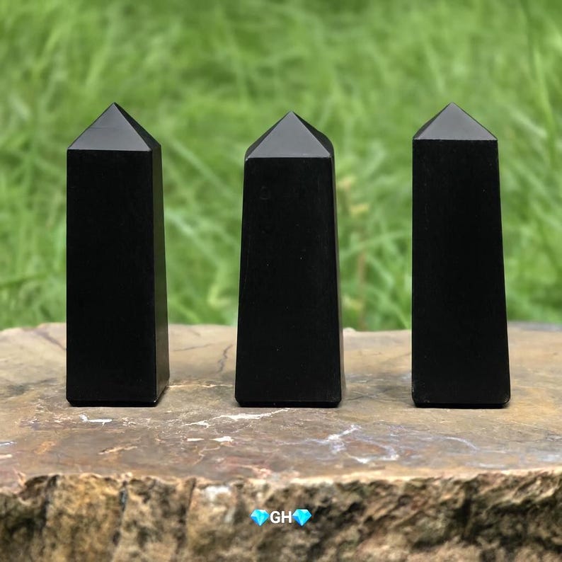 May include: Three polished black obsidian obelisks with a pyramidal top. The obelisks are standing on a stone surface with a green background. The obelisks are approximately 4 inches tall.
