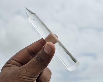 以下が含まれることがあります： 透明で円筒形のクリスタルワンド。先端は尖っており、全体的に滑らかで磨き上げられています。ガラスのような質感で、鋭い円錐形の先端と平らな円形の底面を持ち、表面には光が反射しています。