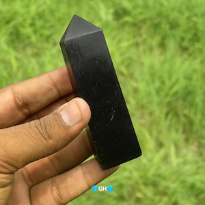 May include: A polished black obelisk-shaped crystal, held in a hand against a blurred green background. The crystal has a flat top and rectangular body. The crystal is approximately 5 inches in length.