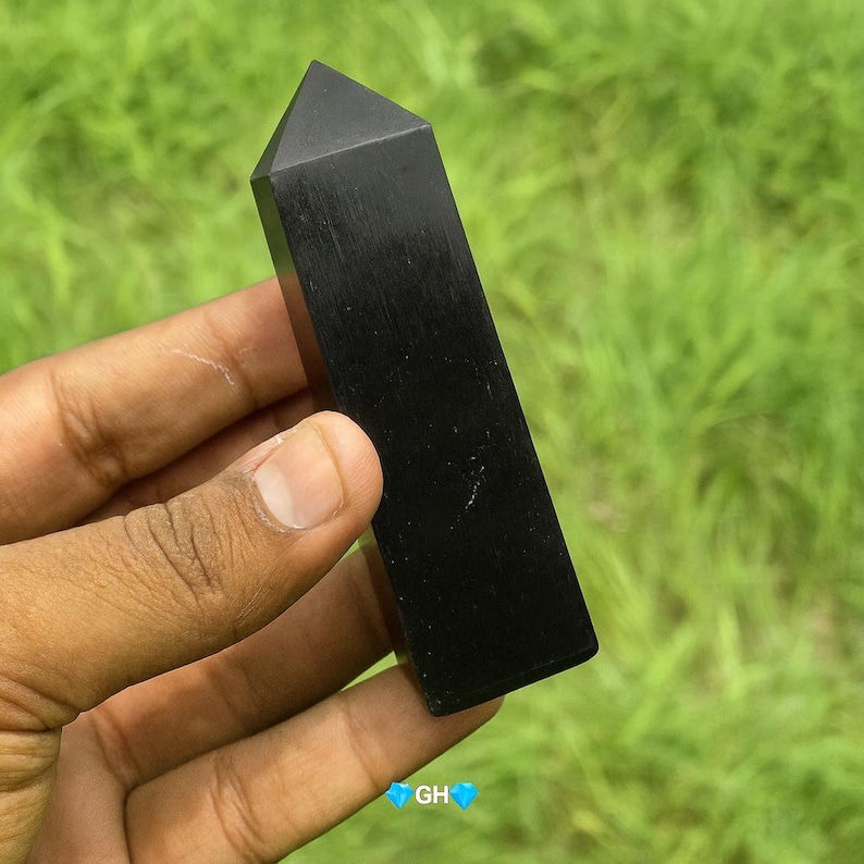 May include: A polished black obelisk-shaped crystal, held in a hand against a blurred green background. The crystal has a flat top and rectangular body. The crystal is approximately 5 inches in length.