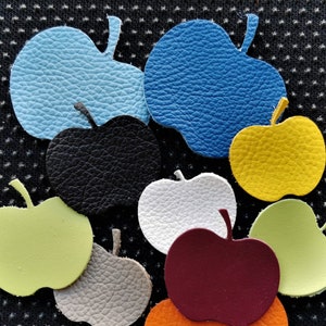 May include: Nine leather apple shapes in various colours, including blue, green, yellow, black, white, brown, and orange. The apples are arranged in a cluster on a black textured surface.
