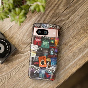 May include: A phone case featuring a collage of Stephen King book covers. The case is wrapped around a phone with a rose gold frame and black camera lenses. The background is a wooden surface.