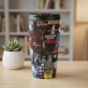 May include: A black travel mug featuring a collage of Stephen King book covers. The mug includes titles like "Misery" and "The Shining." The mug has a black lid and is displayed on a wooden surface.