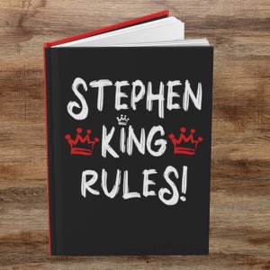 May include: Black notebook with white text that reads "Stephen King Rules!" with two red crowns on either side of the word "King".