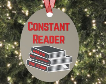 May include: A circular, grey Christmas tree ornament with the words "CONSTANT READER" in red. Below the text are three stacked books with the titles "CARRIE", "PET SEMATARY", and "THE STAND". A red ribbon is attached.