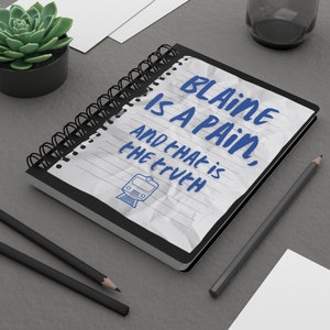 May include: A spiral-bound notebook with the text "BLAINE IS A PAIN, AND THAT IS THE TRUTH" in blue, along with a train illustration. The notebook is open on a gray surface, with pencils and a small succulent plant nearby.