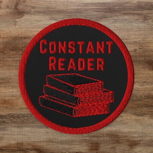 以下が含まれることがあります： 黒い背景と赤い縁取りの刺繍パッチ。パッチには、赤い文字で「CONSTANT READER」と書かれており、その下に同じく赤い3冊の本が重ねて描かれています。パッチは円形で、木製の背景に配置されています。