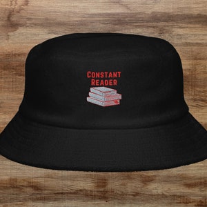 May include: Black bucket hat with the text "CONSTANT READER" embroidered in red and a stack of books in gray.