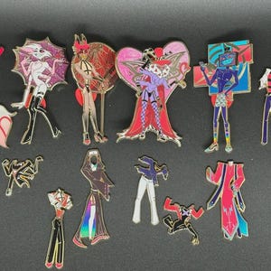 Op de afbeelding: Een verzameling kleurrijke emaille pins met verschillende stripfiguren. De pins tonen gedetailleerde ontwerpen met goudkleurige contouren en levendige kleuren, waaronder roze, rood, paars en blauw. De pins beelden volledige figuren en gestileerde vormen uit.